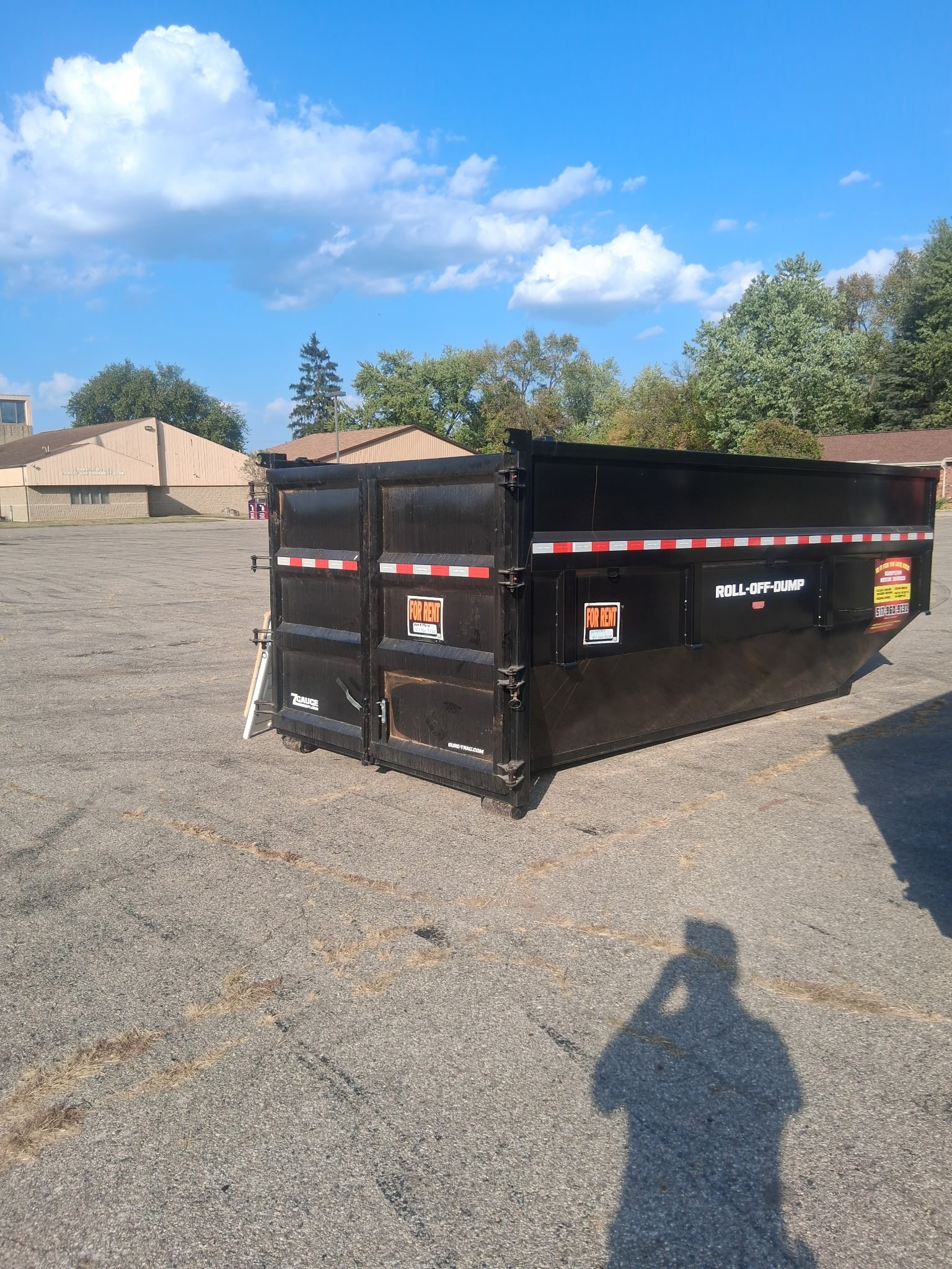 Black roll-off dumpster trailer parked in an industrial lot with buildings and trees in background under blue sky