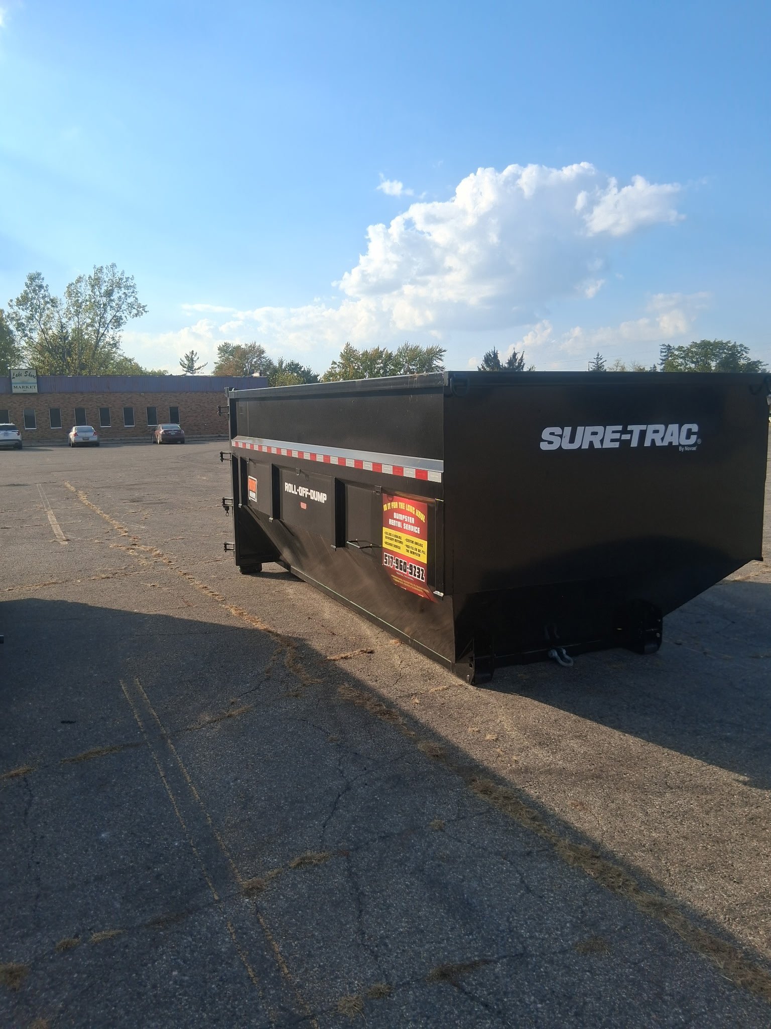 Black Sure-Trac trailer parked in a lot with a commercial building and blue sky with clouds in the background