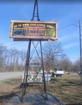 Old roadside sign mounted on metal pole with bare trees and buildings in background against blue sky