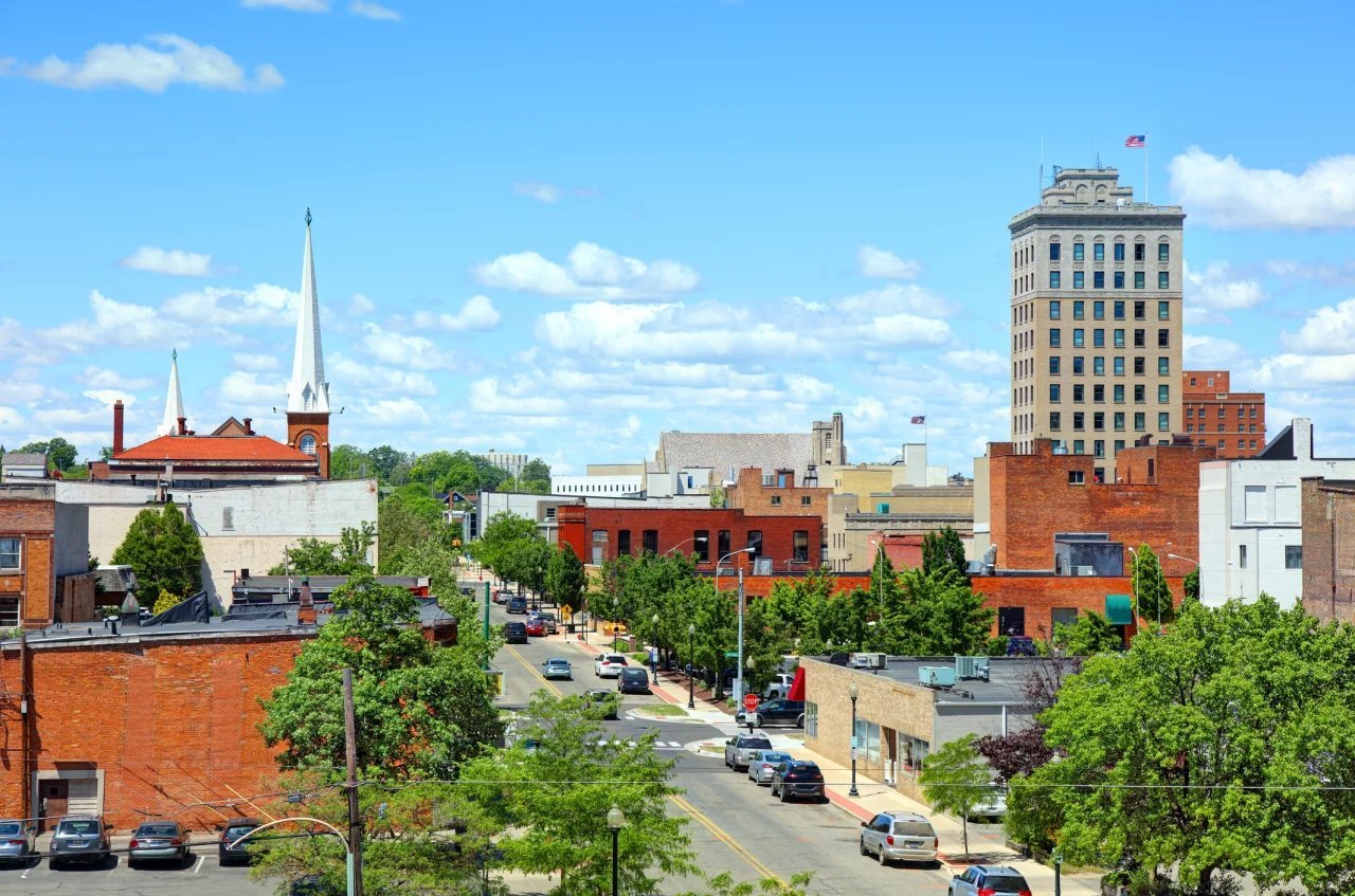 Urban downtown street view with brick buildings, trees, parked cars, and tall office buildings under blue sky with clouds