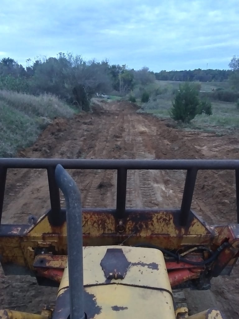 View from a rusty yellow farm vehicle hood of a dirt road through scrubland with trees and fields
